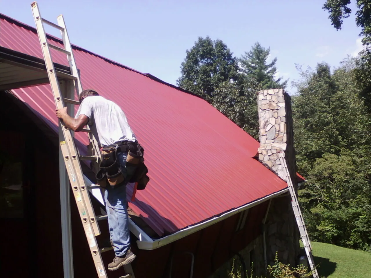 Roofer on a ladder beside a red metal roof and stone chimney — Soffit & Fascia Repair by Roofing Brothers in Aberdeen Proving Ground